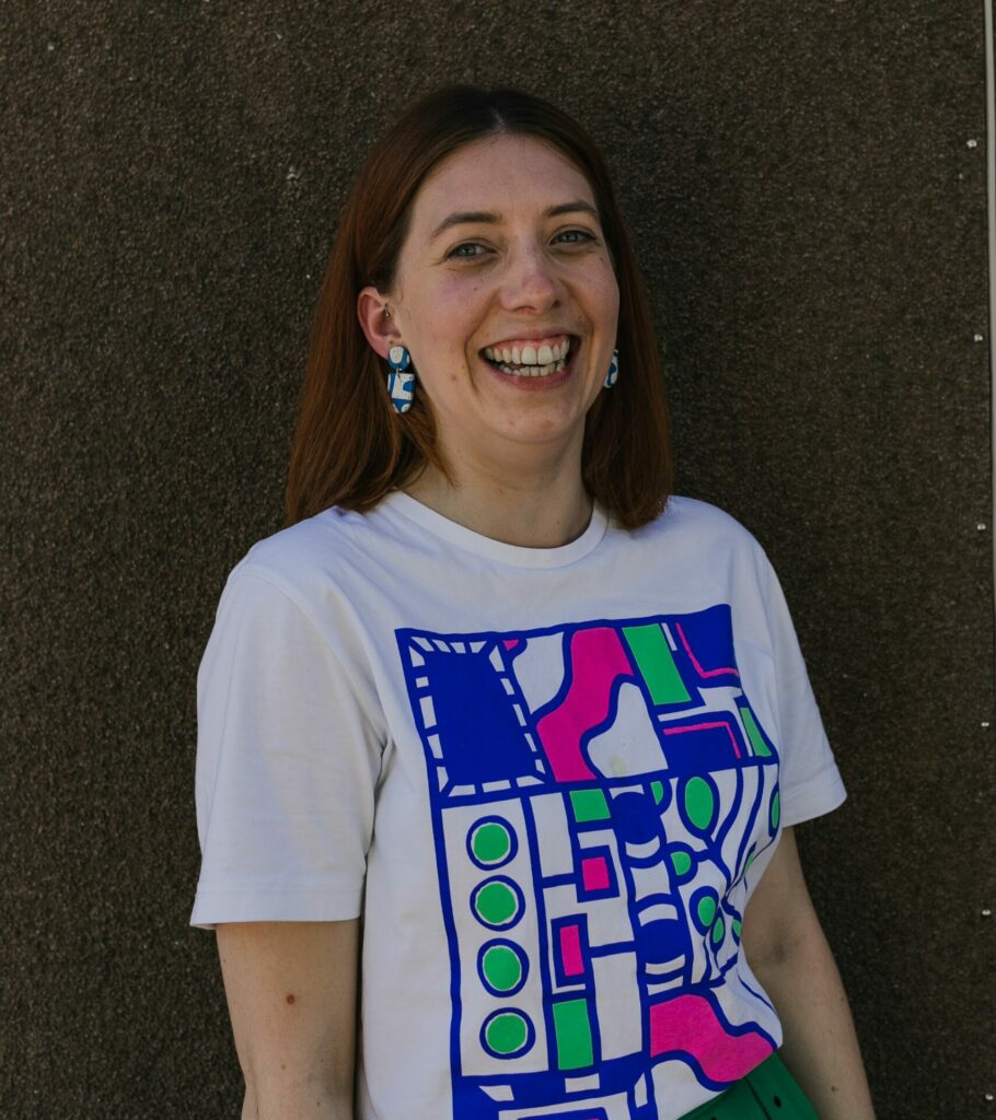 A photo of Becky Jones, Community Producer. Becky is a young white woman with shoulder-length brown hair. She is wearing colourful chunky earrings, a white t-shirt with a brightly coloured pattern, a green skirt and a wide smile.