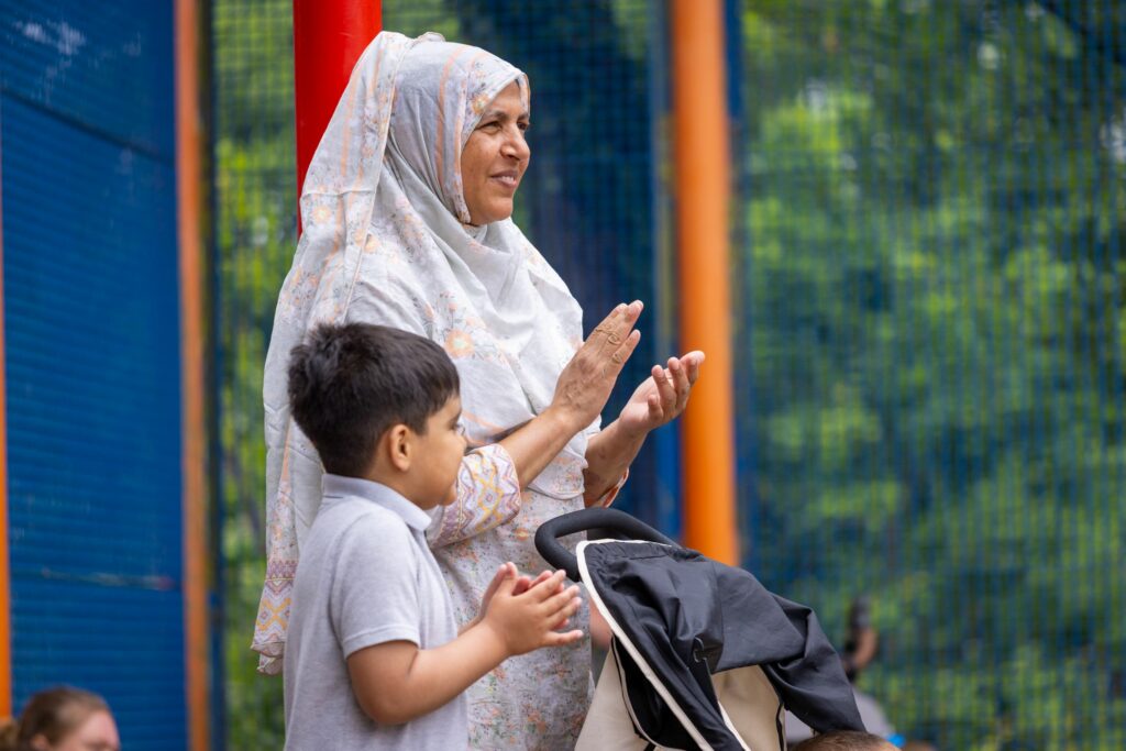 A woman wearing a pale floral hijab stands behind a buggy with a little boy next to her. They both smile and clap their hands.