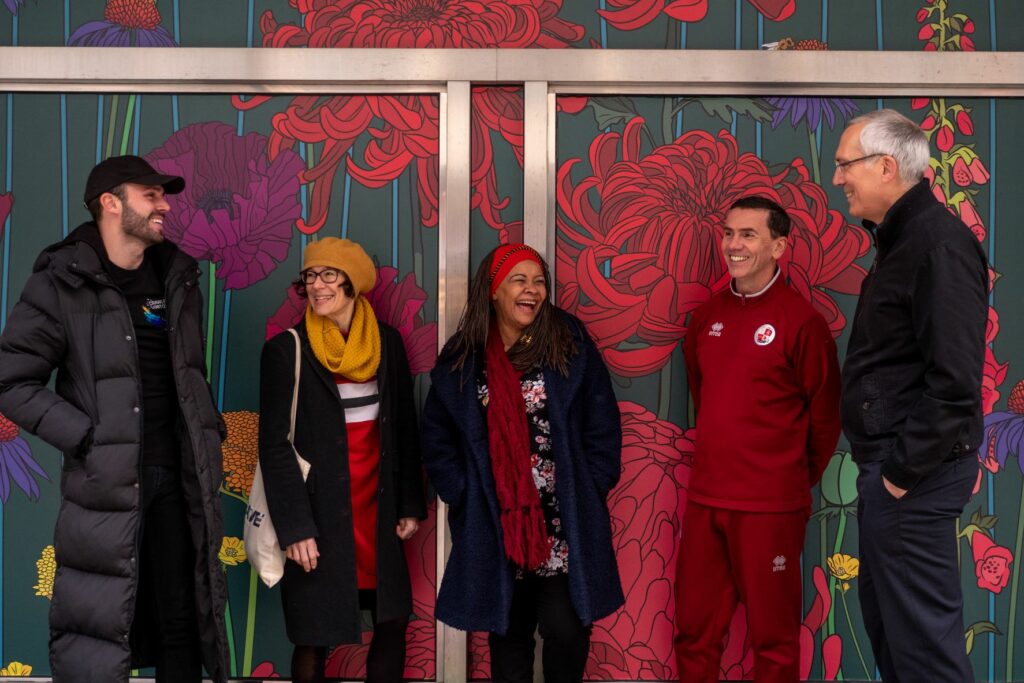 A picture of the Creative Crawley team showing three men and two woman standing laughing and smiling against a wall painted with brightly coloured flowers.