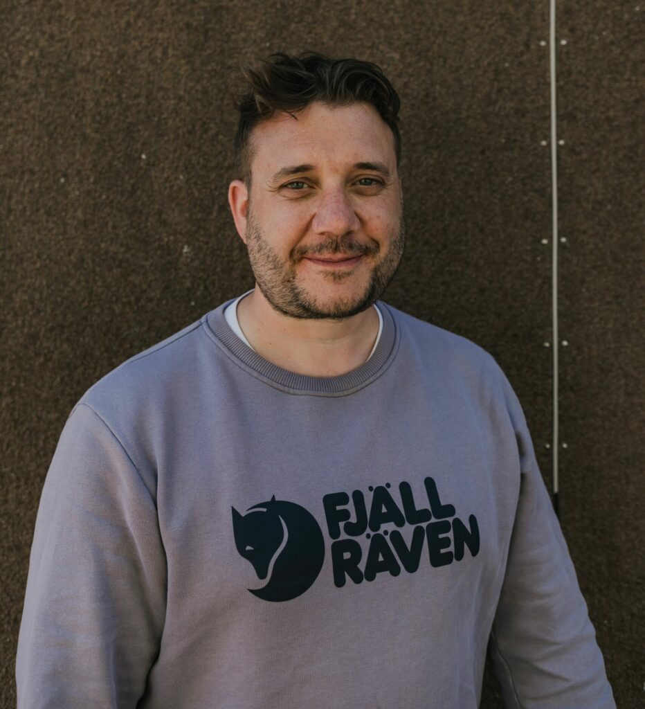 A middle-aged white man with short brown hair and a black and grey short beard stands against a brown background, wearing a grey sweatshirt with a large black logo.