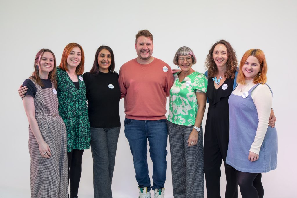 A picture of the Creative Playground team standing in a white backdrop. From left to right: Erin, a young white woman with brown hair wearing black and white check dungarees; Becky, a young white woman with ginger hair wearing a green and black dress; Pav, a middle aged Asian woman with long brown hair wearing a black blouse and grey trousers; Sam, a middle-aged white man with short brown hair and grey stubble, wearing a red sweatshirt and blue denim jeans; Louise, a middle aged white woman with a short bob and glasses wearing a cream and green floral blouse; Sophie, a middle-aged white woman with long curly brown hair wearing a black dress; and Harriet, a young white woman with ginger hair wearing a pale blue dress with black tights.