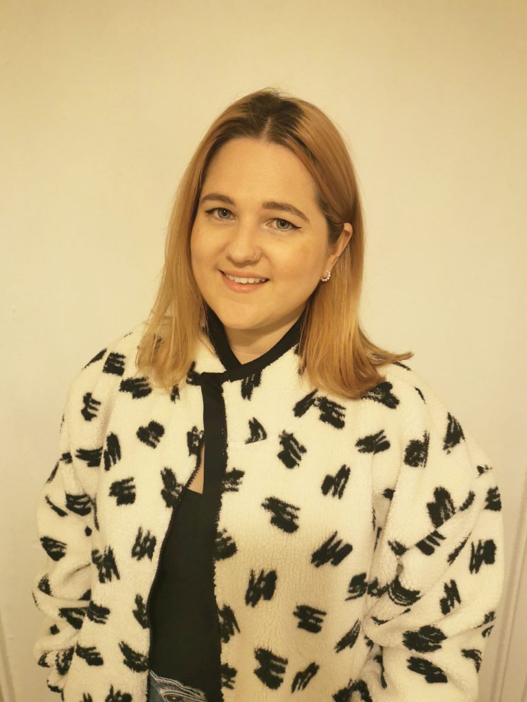 A photo of Harriet McDermott, Communications & Admin Assistant. Harriet is a young white woman with short ginger hair. She is wearing a white fleece with black shapes on and is smiling.