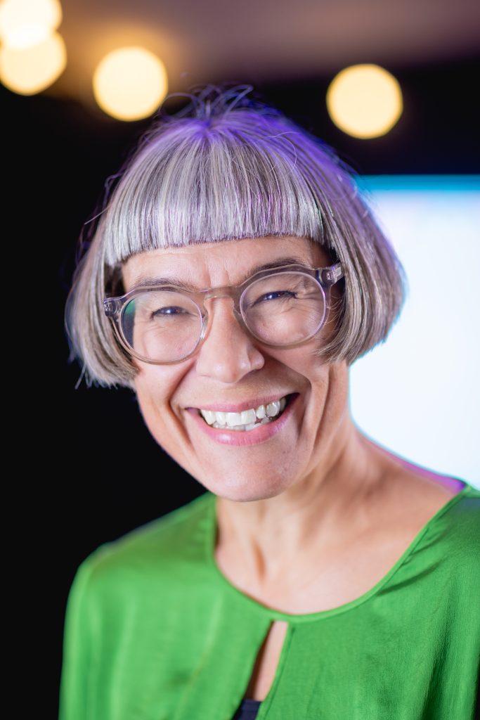 A photo of Louise Blackwell, Co-Director of Creative Playground. Louise is a middle aged white woman with short bobbed grey hair and glasses. She is wearing green blouse and a wide smile.