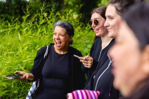 Three women dressed in black casual wear and looking to the left with big smiles on their faces. Two are are holding out their mobile phones. Blurred in the foreground is another woman's face and the background shows bright green, wild bushes.