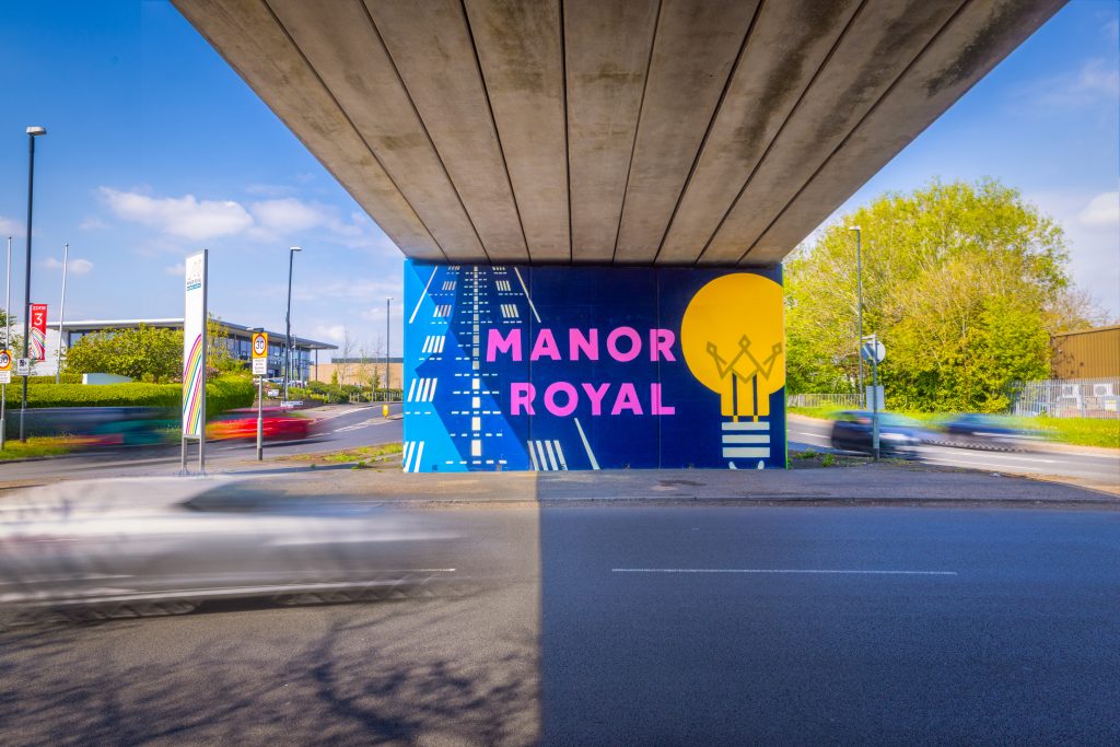 A road with a flyover road over the top. The wall in front has a navy blue background with pink letters reading MANOR ROYAL with a yellow bulb next to the writing. In the background, there are buildings and a tree.